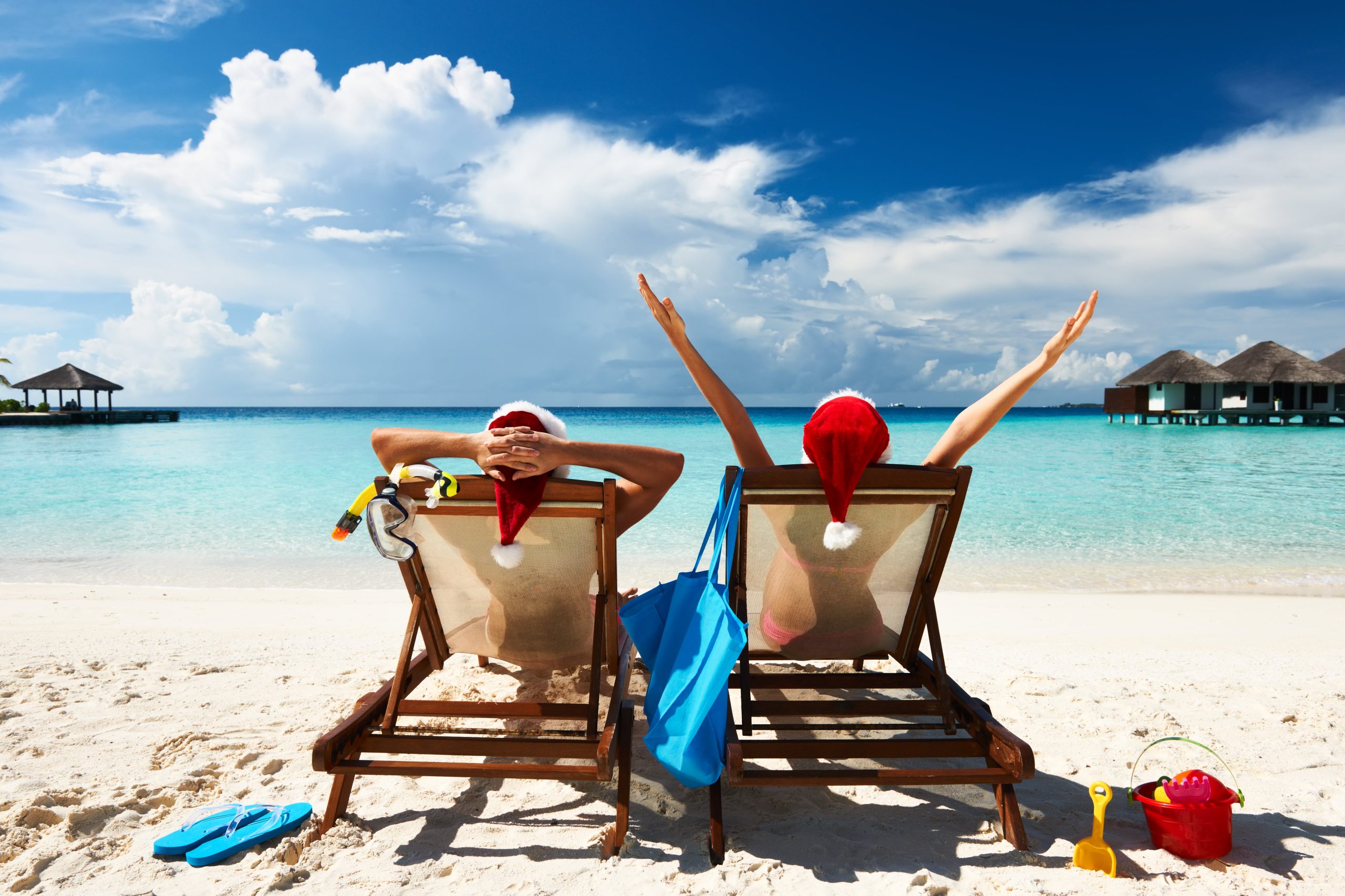 Mentally healthy xmas. Couple sitting on chairs at the beach wearing santa hats, looking out at water. Relaxed.
